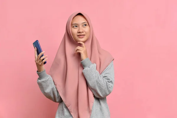 年轻的亚洲女孩手里拿着智能手机 望着别处 用粉色背景与复制空间隔离 — 图库照片