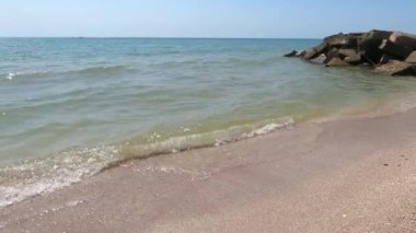 Turkuaz deniz dalgaları kıyı kayaları üzerinde kırılır ve yoğun bir beyaz köpük oluşur. Açık mavi gökyüzü. Beyaz dalgalar kumlu sahilde kırılıyor denizdeki kayalıklarda.