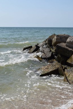 Turkuaz deniz dalgaları kıyı kayaları üzerinde kırılır ve yoğun bir beyaz köpük oluşur. Açık mavi gökyüzü. Beyaz dalgalar kumlu sahilde kırılıyor denizdeki kayalıklarda.