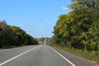 Kırsal kesimde otoyol, yol kenarı ve gri asfalt, yeşil ağaçlar ve tarlalar, yaz gününde mavi gökyüzü. Yazın ormanda boş asfalt yolu olan bir manzara. Seyahat