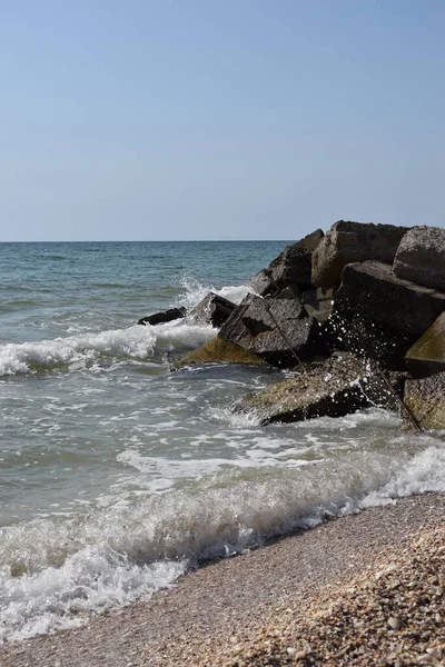 Turkuaz deniz dalgaları kıyı kayaları üzerinde kırılır ve yoğun bir beyaz köpük oluşur. Açık mavi gökyüzü. Beyaz dalgalar kumlu sahilde kırılıyor denizdeki kayalıklarda.
