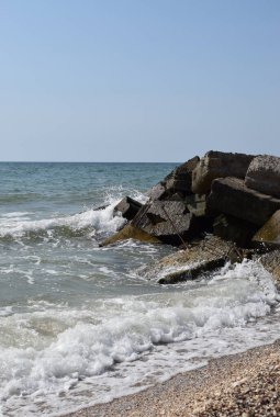 Turkuaz deniz dalgaları kıyı kayaları üzerinde kırılır ve yoğun bir beyaz köpük oluşur. Açık mavi gökyüzü. Beyaz dalgalar kumlu sahilde kırılıyor denizdeki kayalıklarda.
