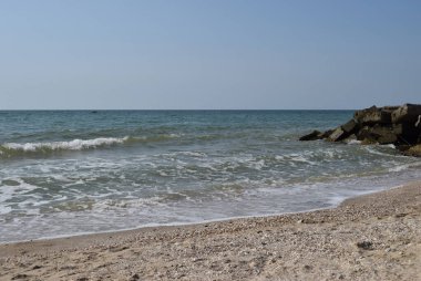 Turkuaz deniz dalgaları kıyı kayaları üzerinde kırılır ve yoğun bir beyaz köpük oluşur. Açık mavi gökyüzü. Beyaz dalgalar kumlu sahilde kırılıyor denizdeki kayalıklarda.