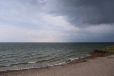 Bulutlu gri bir gökyüzü bu sahildeki beyaz köpüklü deniz mavisi sularını örtüyor. Kara fırtınalı bulutlu gri gökyüzü. Bir günde alçak bulutlu gökyüzünün altındaki kumsal.