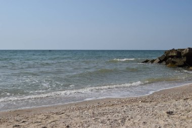 Turkuaz deniz dalgaları kıyı kayaları üzerinde kırılır ve yoğun bir beyaz köpük oluşur. Açık mavi gökyüzü. Beyaz dalgalar kumlu sahilde kırılıyor denizdeki kayalıklarda.