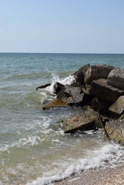 Turkuaz deniz dalgaları kıyı kayaları üzerinde kırılır ve yoğun bir beyaz köpük oluşur. Açık mavi gökyüzü. Beyaz dalgalar kumlu sahilde kırılıyor denizdeki kayalıklarda.