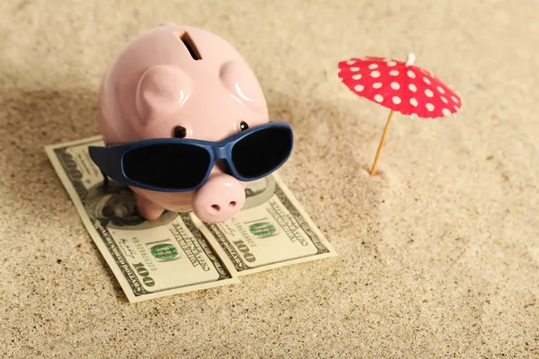 Summer Piggy Bank em pé na toalha de greenback cem dólares com óculos de sol na praia e guarda-sol vermelho — Fotografia de Stock