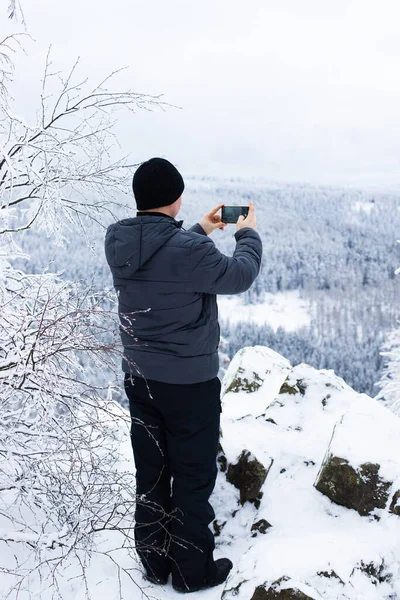 Un uomo in inverno in un parco o foresta fa una foto su un telefono cellulare. Fotografia mobile, passeggiate nel bosco. Paesaggio invernale. Orientamento verticale — Foto Stock