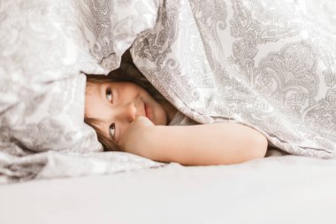 4 yaşında tatlı bir çocuk battaniyenin altından dışarı bakıyor. Evde sabah eğlencesi. Hayatın şimdiki anları. Çocuklarla yatakta oynamak.