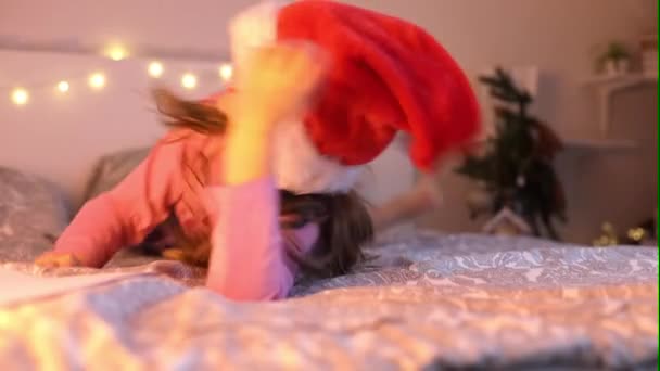 Egy aranyos kislány szánkókalapban kényezteti az ágyon karácsony este. Gyerekkori álmok. Újévi ünnepek. A gyerekek várják a Mikulást. — Stock videók
