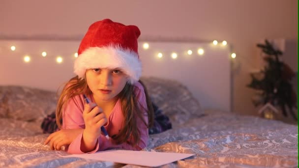 Una linda chica en un sombrero sana escribe una carta a Santa Claus para Navidad con deseos y sueños sobre regalos. Sueños de infancia. Vacaciones de Año Nuevo — Vídeo de stock