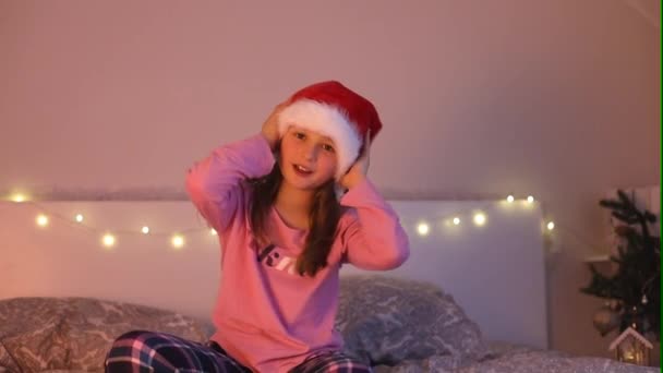 Uma menina bonita em um chapéu de Papai Noel está brincando na cama e puxa o chapéu sobre sua cabeça. À espera do Ano Novo e do Natal. Feriados infantis. Milagre — Vídeo de Stock