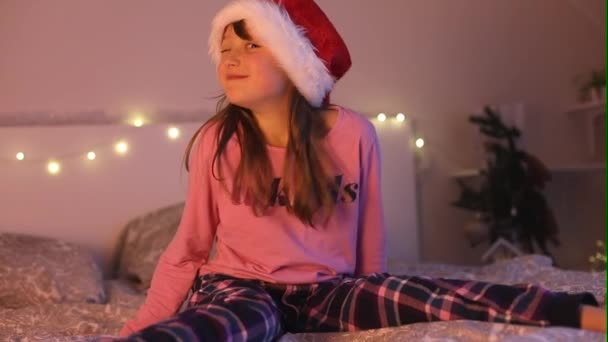 Καλά Χριστούγεννα και Καλή Χρονιά. Ένα κορίτσι με καπέλο 'γιου Βασίλη παίζει στο κρεβάτι, κλείνει το μάτι και πέφτει στο κρεβάτι. Παιδικές διακοπές. — Αρχείο Βίντεο
