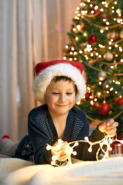 Noel ve Yeni Yıl. Süslü bir Noel ağacının yanında çelenklerle oynayan çocuk. Şenlik büyüsü