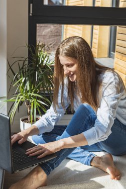 Genç bir kız pencerenin kenarında dizüstü bilgisayarın başında oturuyor ve yazıyor. Çevrimiçi iletişim. Uzaktan kumanda. Bağımsız