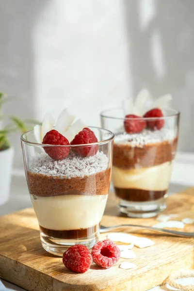 Sobremesa de mousse de três camadas feita de chocolate e baunilha, decorada com framboesas frescas e flocos de coco em copos na mesa. Brunch, sobremesa para os amantes — Fotografia de Stock