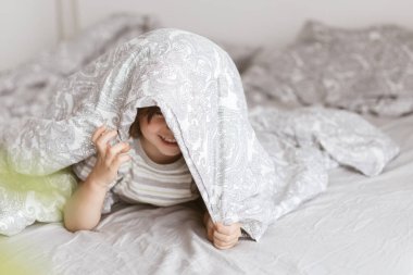 Sevimli bir çocuk, yeni yürümeye başlayan, ailesinin yatağında eğlenen, battaniyeyle kendini örten ve gülen. Evde oyunlar