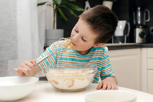 Mignon garçon lèche un fouet avec de la pâte à gâteau dans la cuisine à la maison. Du temps avec les enfants à la maison. Petite assistante. Les enfants préparent la nourriture — Photo