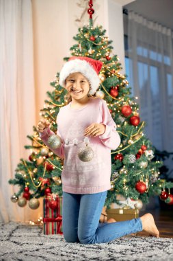 Mutlu Noeller ve yeni yıl tatilleri. Çekici küçük bir kız Noel ağacını süslüyor ve Noel süslemeleriyle oynuyor. Dikey fotoğraf. Hayattan anlar.