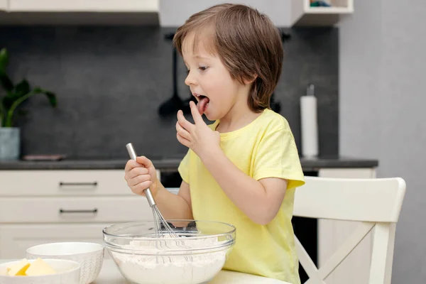 Un petit garçon pétrit la pâte avec un fouet et la goûte. Les enfants préparent un gâteau. Petit chef et assistant — Photo