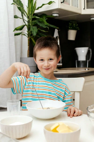 Un petit garçon pétrit la pâte avec un fouet et la goûte. Les enfants préparent un gâteau. Petit chef et assistant — Photo