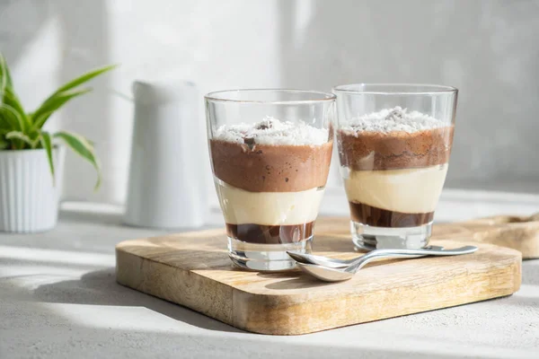 Lahodná třívrstvá pěna, vanilka, čokoláda, krémový dezert ve sklenici v jasných slunečních paprscích. — Stock fotografie