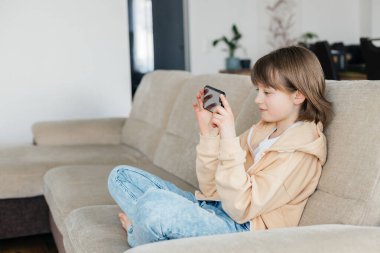 Öğrenci kız kanepede oturuyor ve cep telefonuyla oynuyor ya da arkadaşlarıyla sohbet ediyor. Çocuklar teknolojiyi kullanır. Modern aletler ve gençler..