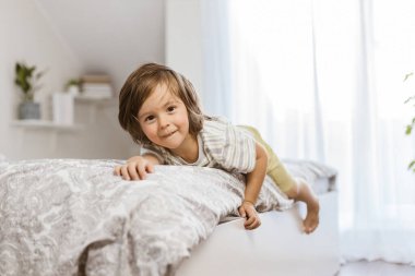 Sevimli, mutlu bir çocuk yatak odasındaki yatağın kenarına uzanıp kameraya bakıyor. Çocuklar evde oynuyor. Çocuklukta kahkaha ve eğlence.
