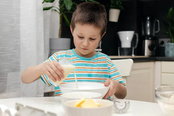 Le garçon prépare la pâte pour le biscuit ou la cuisson dans la cuisine. L'enfant ajoute du lait dans le bol. Petit chef et assistant dans la cuisine. — Photo