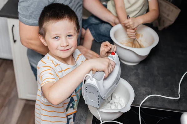 The boy helps to knead the dough with a mixer in a bowl in the kitchen at home. Satisfied child looks at the camera and smiles while cooking. — Stock Photo, Image