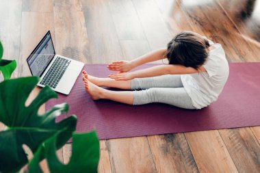 Spor yapan sevimli küçük kız, evde esneme hareketleri yapıyor. Çocuk evde internetten yoga öğreniyor. Çocuk sporları ve evdeki aktiviteler. Çocuklar için yoga pozları.