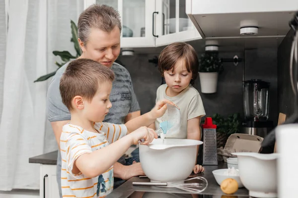 Caring dad is cooking in the kitchen with his two sons. Boys and dad prepare pie dough, muffins, pancakes for breakfast at home. Family time together.