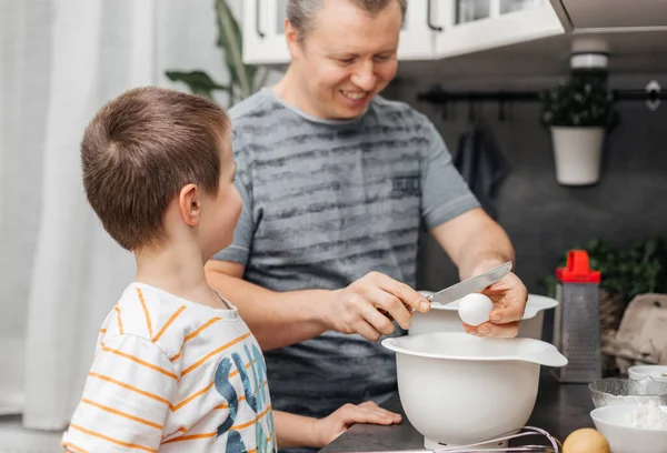 Dad and son are cooking in the kitchen. Dad teaches to break the childs eggs and cook the dough. Happy time of children and parents together at home