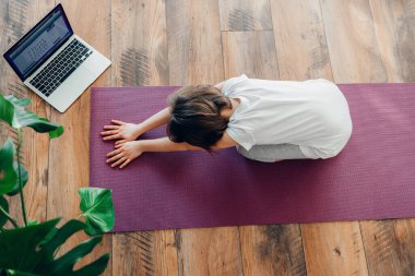 Kız bir spor paspasına oturuyor ve yoga yapıyor. Evde ve internette çocuklar ve yetişkinler için yoga. Bir çocuğun spor yaşam tarzı.