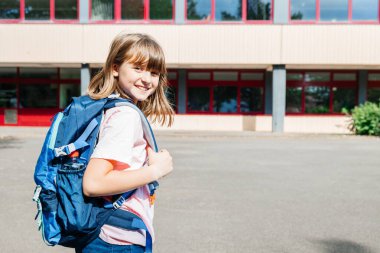 Portrait of a happy schoolgirl girl with a backpack on her back against the school. Children go to school. Beginning or end of lessons and school year.