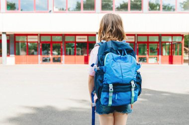 A teenage girl with a school backpack on her back stands in front of the school and looks at her. Beginning of the school year for students. Back to school.
