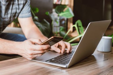 Evde dizüstü bilgisayarın yanındaki bir adamın elindeki telefonun yakın plan görüntüsü. İnternette çalışıyorum. Cihazları kullanarak online alışveriş ve ödeme