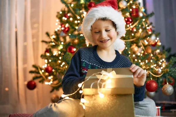 A boy in a santa hat opens a gift box next to a garlanded Christmas tree. Gifts for children. Dreams Come True.
