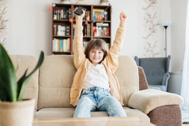 A teenage girl sits on a sofa and joyfully waves her hands up. A child with a mobile phone in his hands. Communication of children online.
