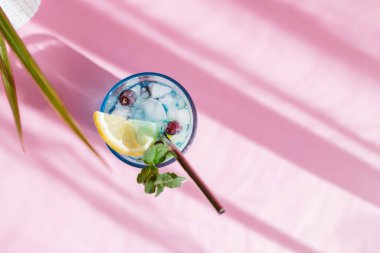 Top view of refreshing blue curacao cocktail on pink background garnished with mint and lemon slice. Colorful summer drinks with fresh berries and shadows.