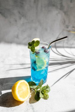 Refreshing blue curacao cocktail garnished with mint and a slice of lemon. Colorful summer drinks with fresh berries, bright sun and shadows.