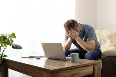 Young pensive male businessman working at home on a laptop. The freelancer sits on a chair and holds his hands behind his head. Work stress.