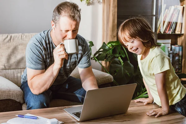 A multitasking young father tries to work from home with his son. A man checks his mail on a laptop and drinks coffee, while a small child looks at the screen. Freelance.