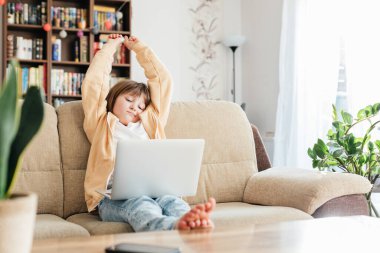 The girl sits on the couch in front of a laptop and stretches from fatigue. Home distance learning for children. Schoolchildren life concept. Children use technology.