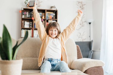 Evdeki çocuklar için internet üzerinden öğrenim. Kız bir dizüstü bilgisayarın önünde kanepede oturur ve zafer işareti olarak ellerini kaldırır. Çocuklar çevrimiçi iletişim. 