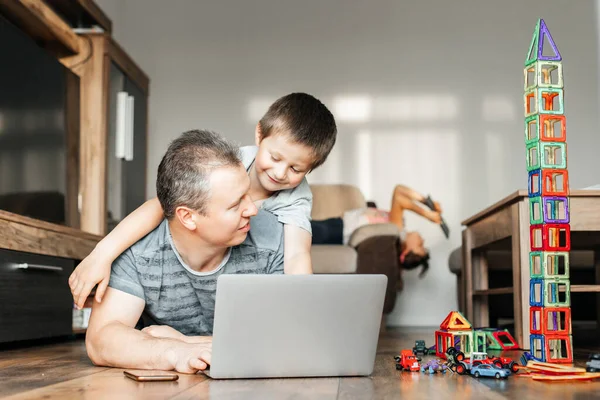 Dad works at home on a laptop and plays with his son at the same time. The child hugs dad and interferes with work. Children and remote work from home.