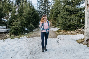 A young beautiful girl walks in the forest with a hiking backpack. Happy female traveler enjoying nature, looking at camera and smiling.