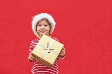 Charming boy in a santa claus hat and a gift box in his hands on a red background with space for text. New Year and Christmas gifts and sales. 