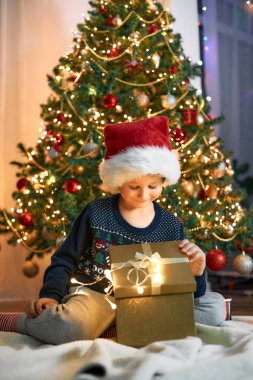 A charming boy in a santa claus hat sits next to the Christmas tree and opens a box with a gift. Christmas and New Year.