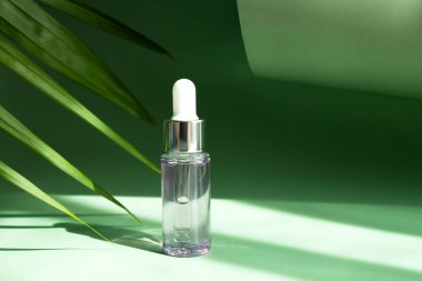 Parlak güneş ışınlarında palmiye yaprağının yanında pipeti olan şeffaf bir kozmetik şişe..